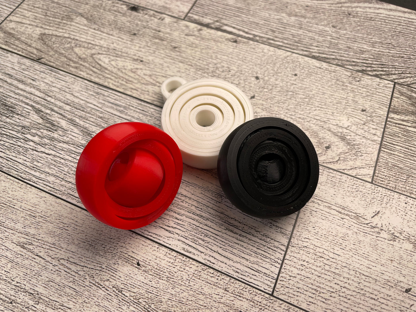 Three small (about 1.5 inches in diameter) 3D printed gyro fidgets on a wood background. Each fidget has four nested circles that can be spun and twirled. The fidgets are printed in red, white and black filament. The white fidget has a keyhole for attaching it to a keychain.