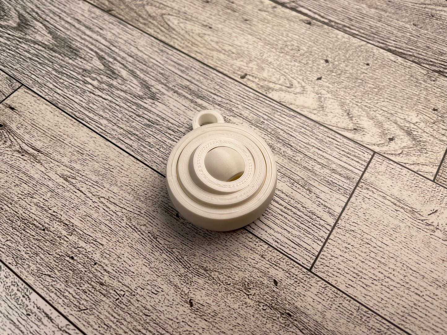 A small 3D printed gyro fidget that is about 1.5 inches in diameter on a wood background. It has four nested circles that can be spun and twirled. It is printed in white filament and has a keyhole for attaching it to a keychain.