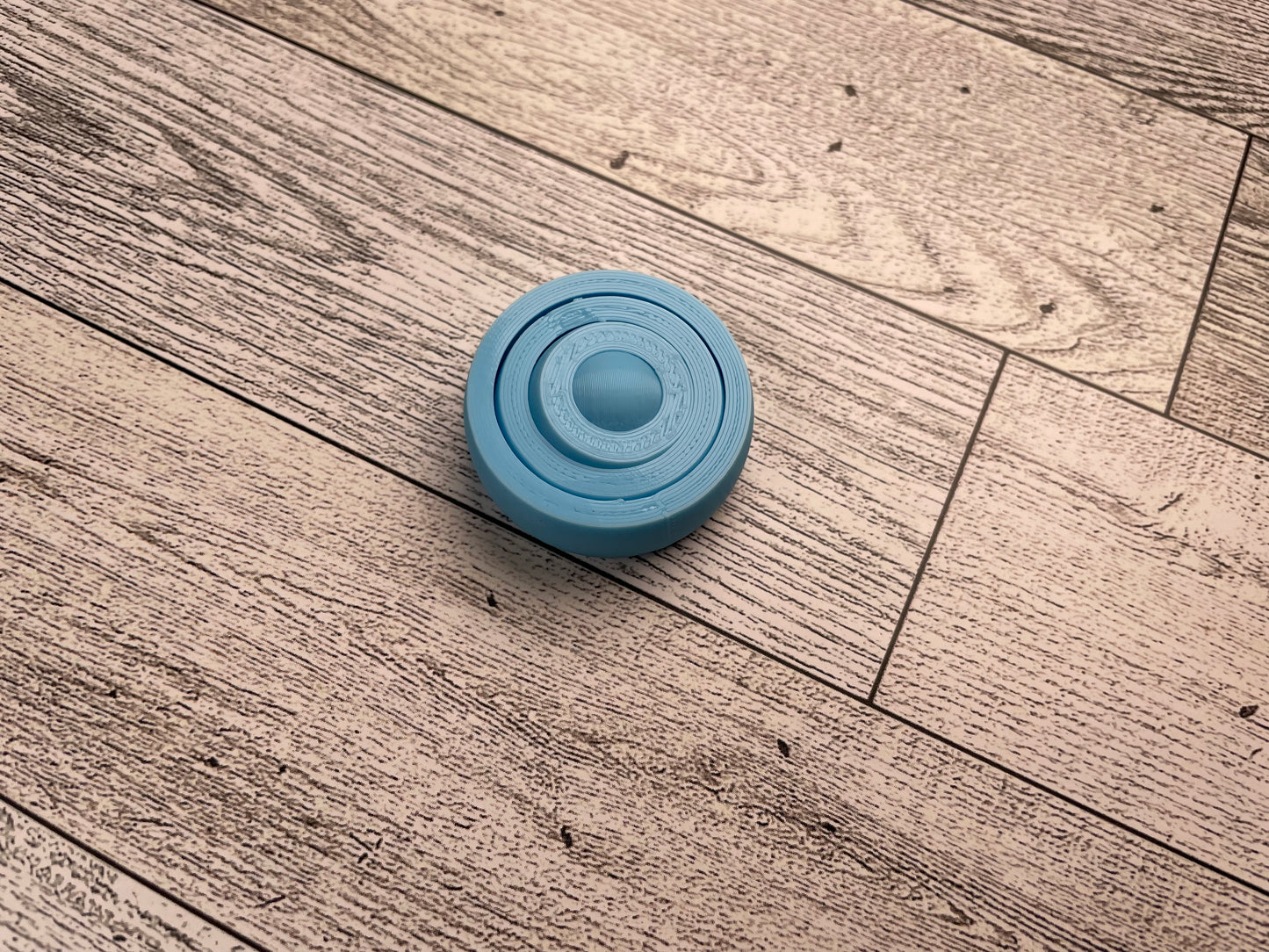A small 3D printed gyro fidget that is about 1.5 inches in diameter on a wood background. It has four nested circles that can be spun and twirled. It is printed in light blue filament.