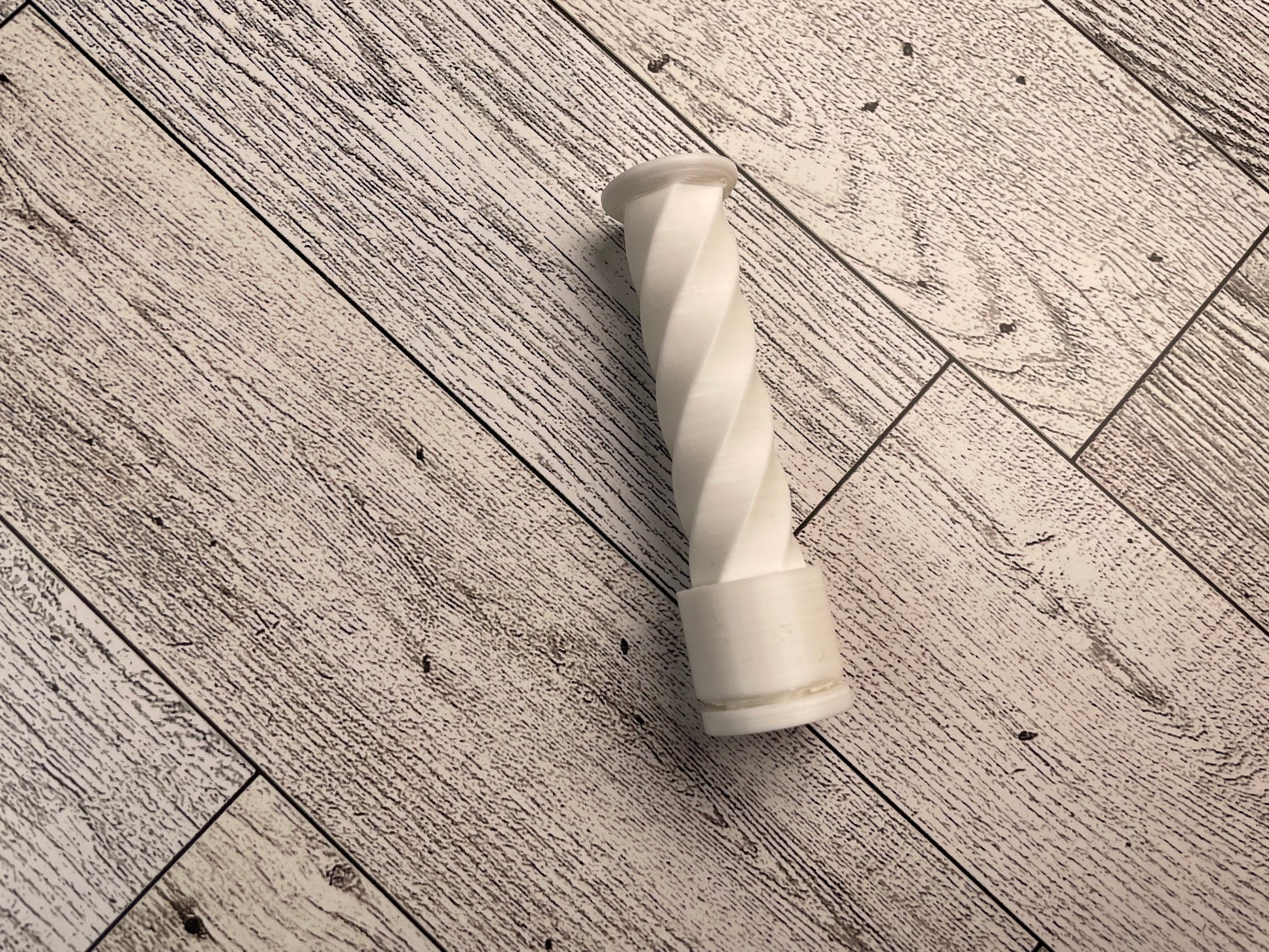 One white spiral fidget on a wood background. The tower is about 3 inches tall and an inch wide and looks like it is twised. There is a cylinder that can move up and down the tower as it spins.