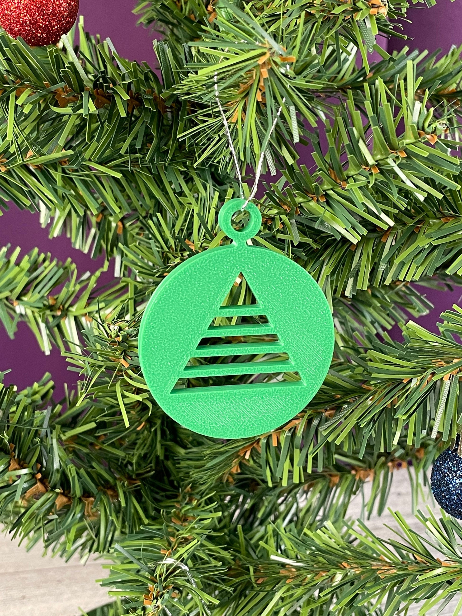 A close up of an ornament printed in green filament hung on a christmas tree with a purple background, it is a circle with a triangular christmas tree cut out of the circle.