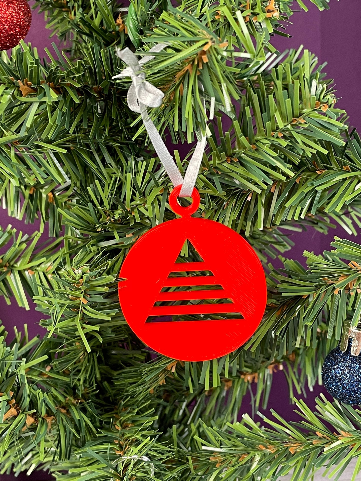 A close up of an ornament printed in red filament hung on a christmas tree with a purple background, it is a circle with a triangular christmas tree cut out of the circle.