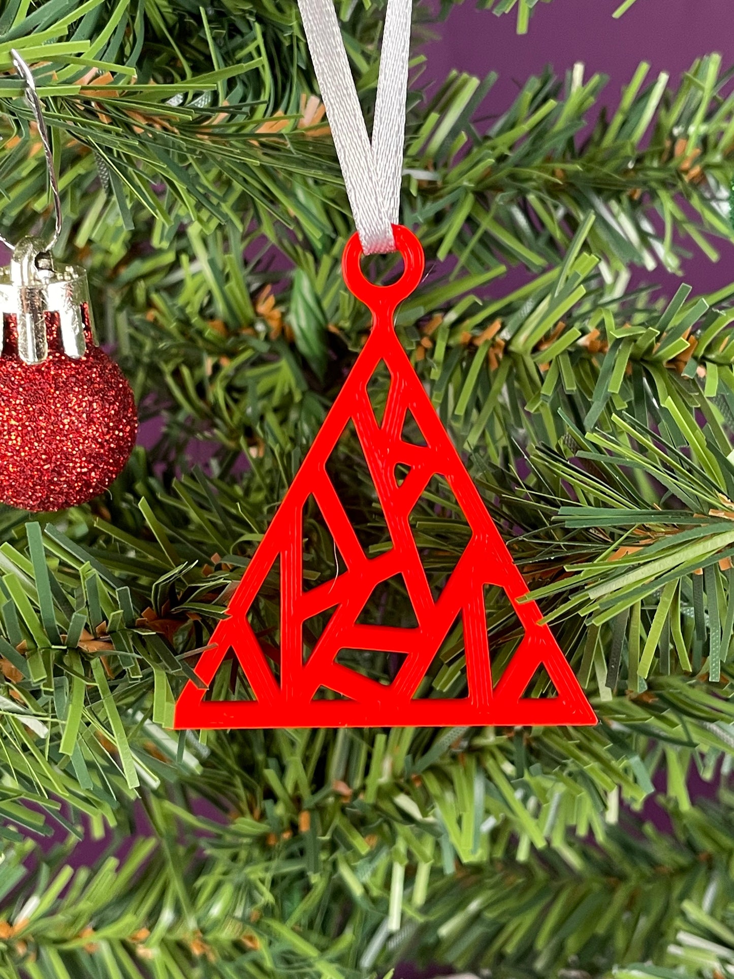 A close up of a fractal tree ornament printed in red filament hung on a christmas tree with a purple background.