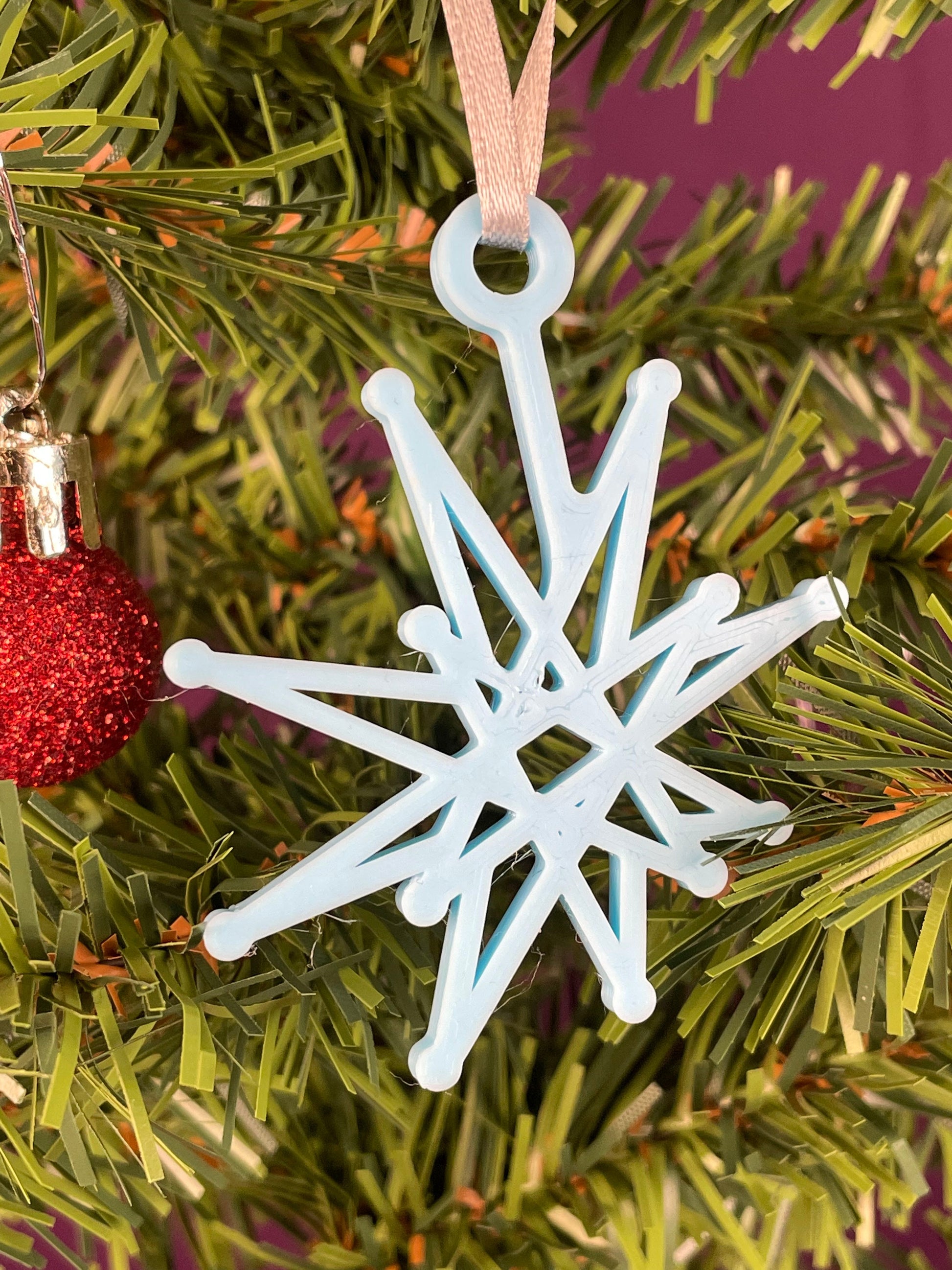 A close up of a fractal starburst ornament printed in light blue filament hung on a christmas tree with a purple background.