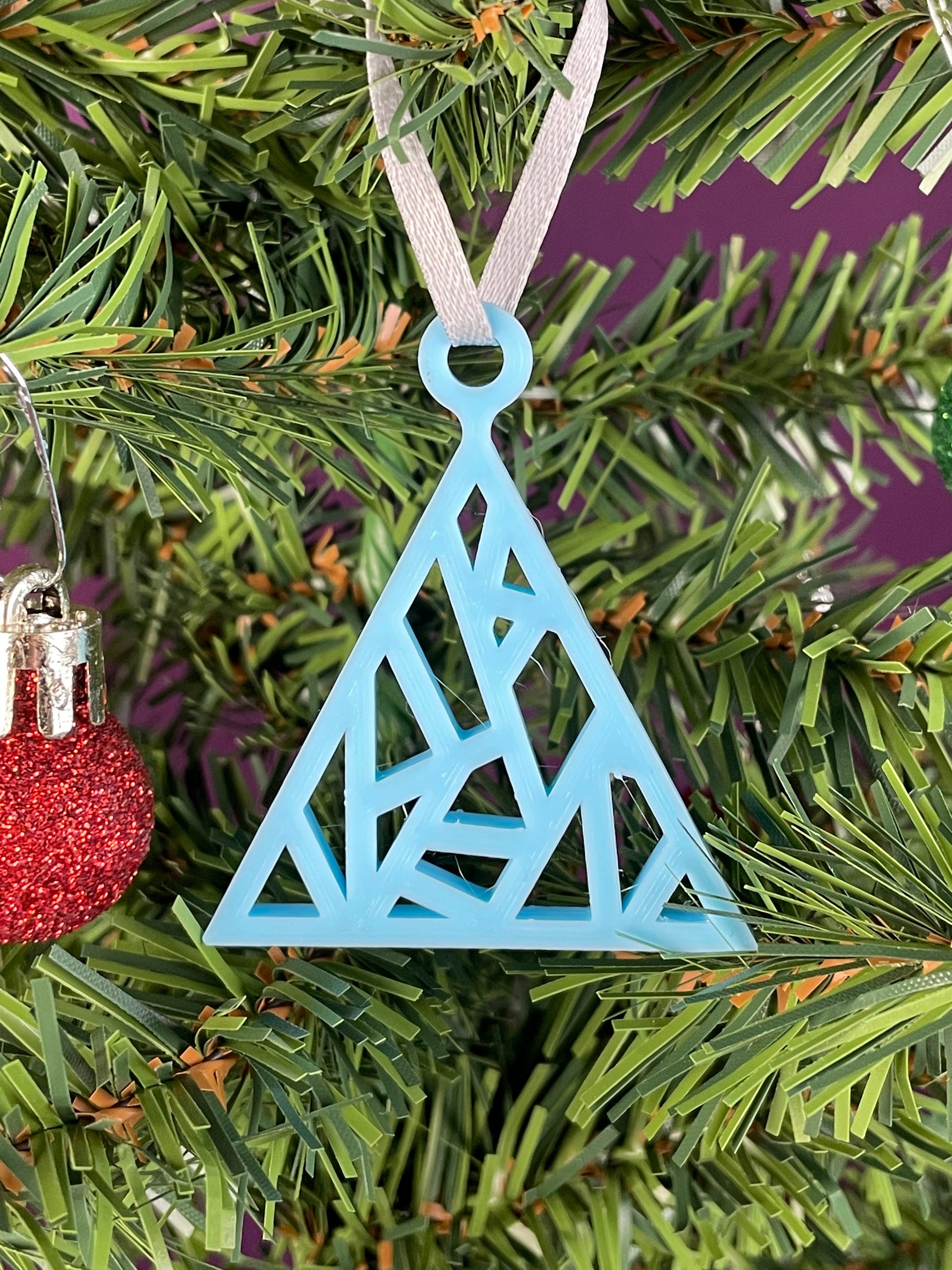 A close up of a fractal tree ornament printed in light blue filament hung on a christmas tree with a purple background.