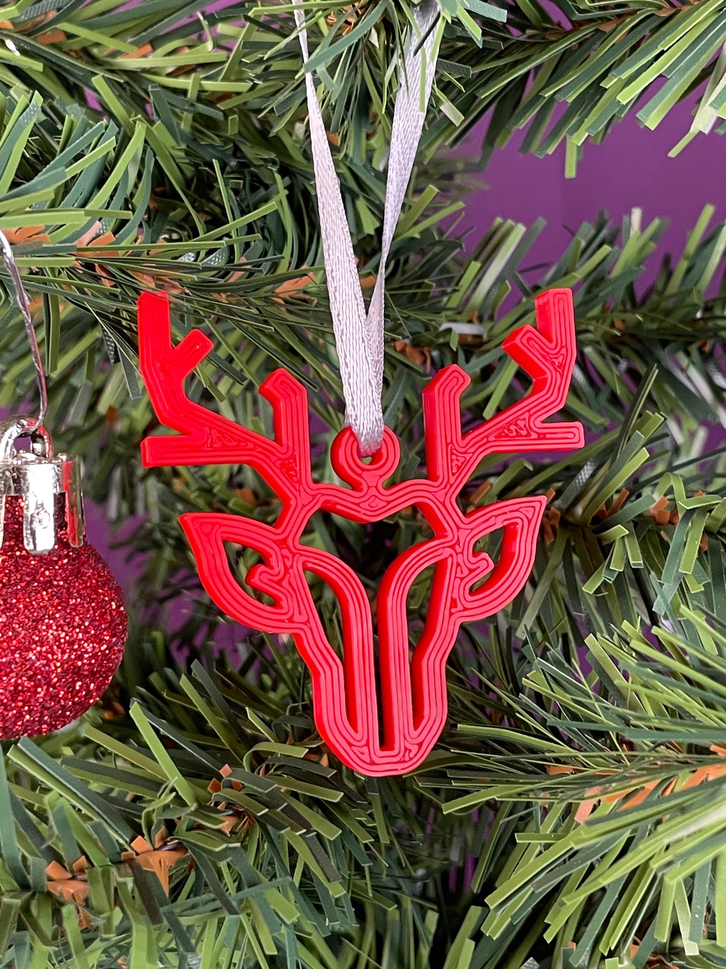 A close up of a modern reindeer ornament printed in red filament hung on a christmas tree with a purple background.