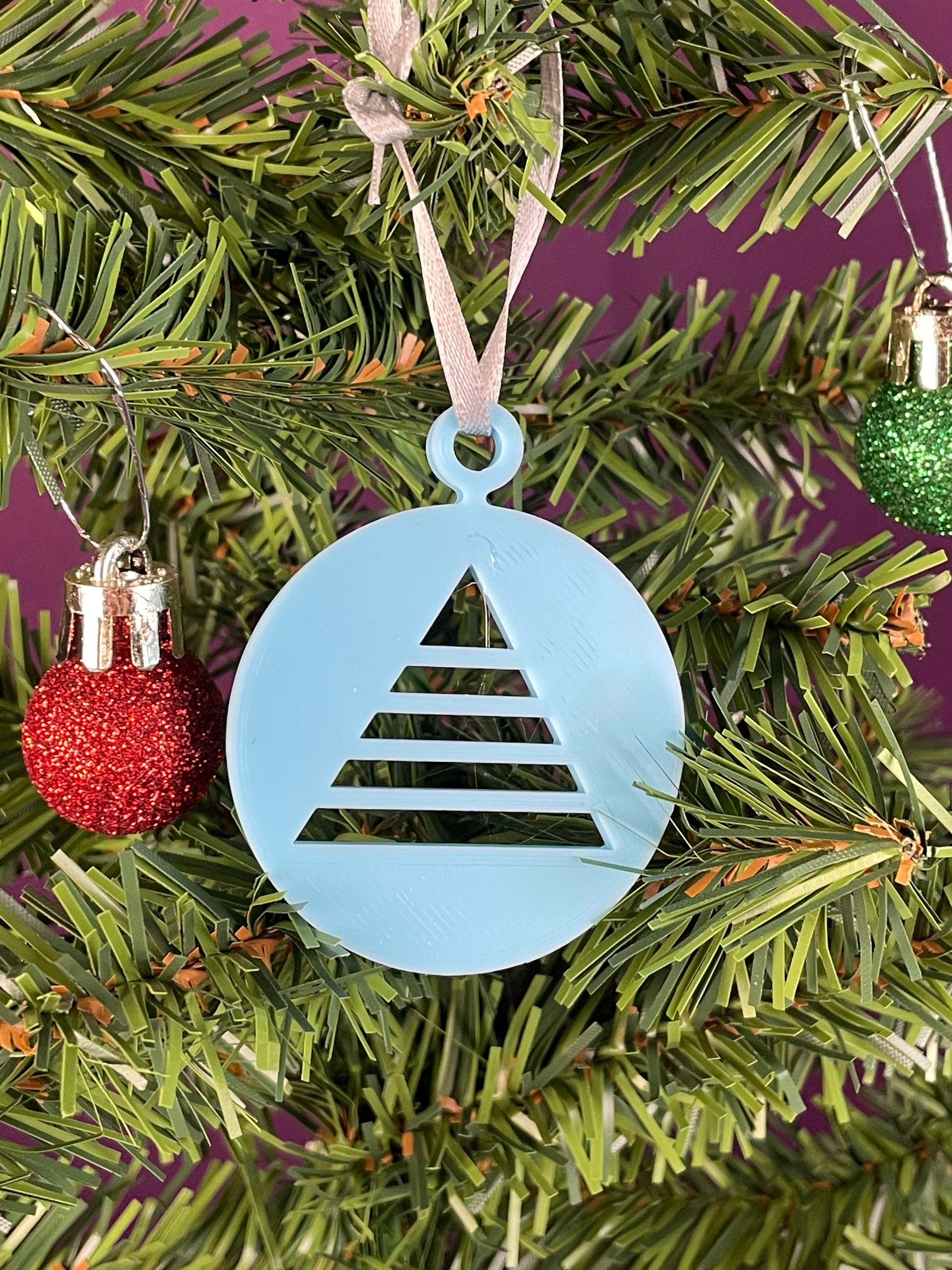 A close up of an ornament printed in light blue filament hung on a christmas tree with a purple background, it is a circle with a triangular christmas tree cut out of the circle.