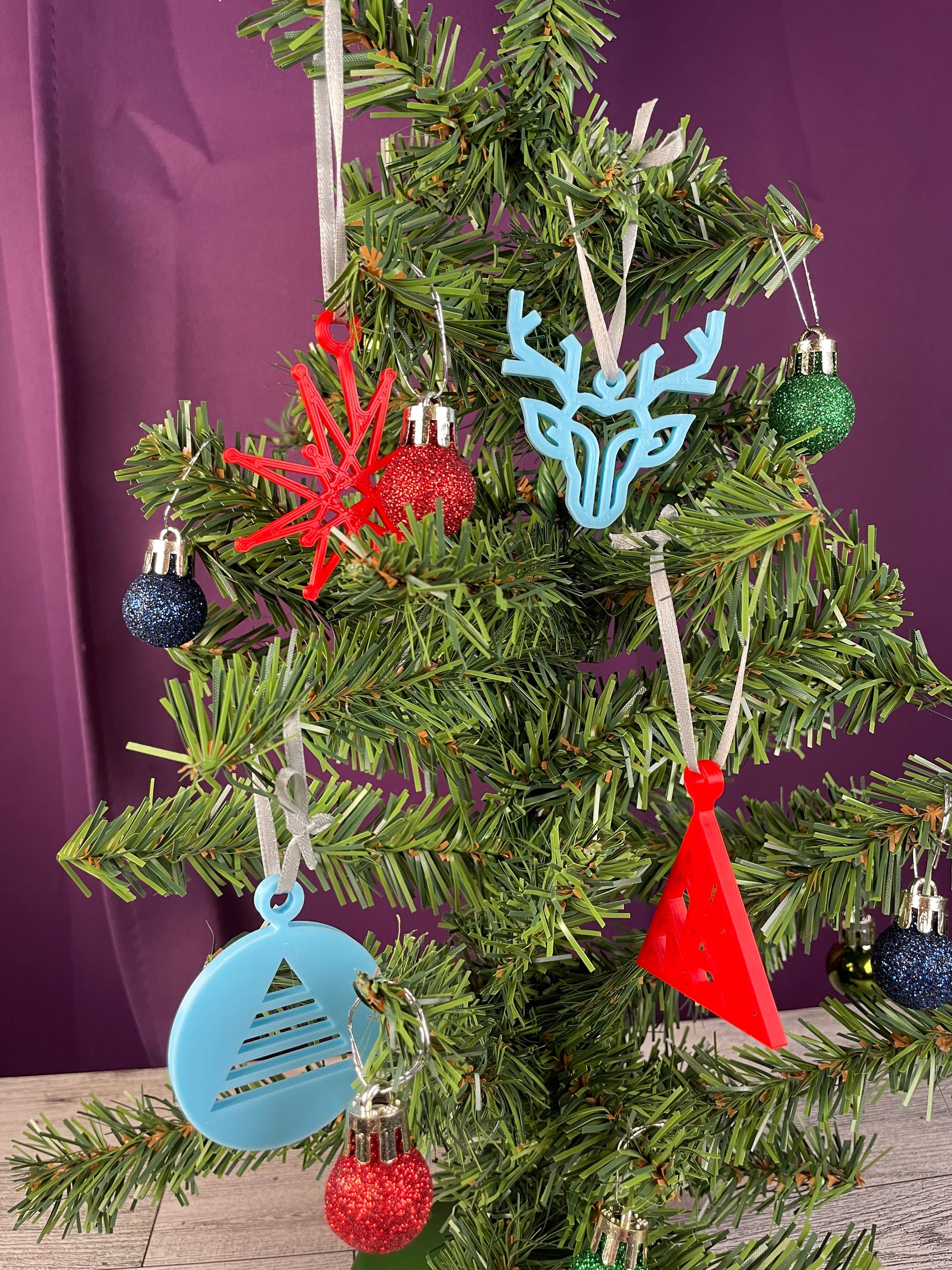 Ornaments on a christmas tree on a wood surface and a purple curtain backdrop. Six ornaments are small sparkly balls in blue, red and green. The other ornaments are a fractal star in red with a light gray ribbon. A light blue modern reindeer with a light gray ribbon. A red fractal christmas tree with a light gray ribbon. The last ornament is a light blue circle with a triangular christmas tree cut out of the circle.